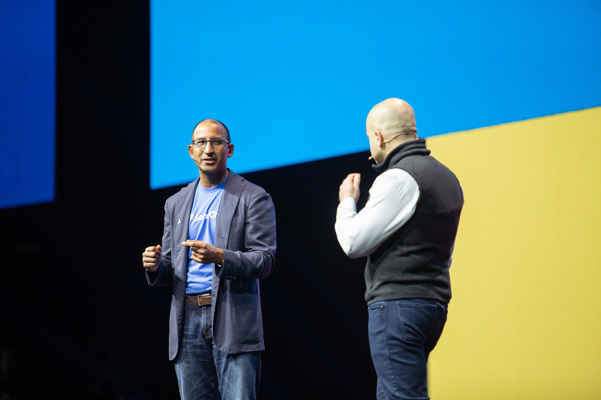 Two executives in conversation on stage with bold blue and yellow split backdrop